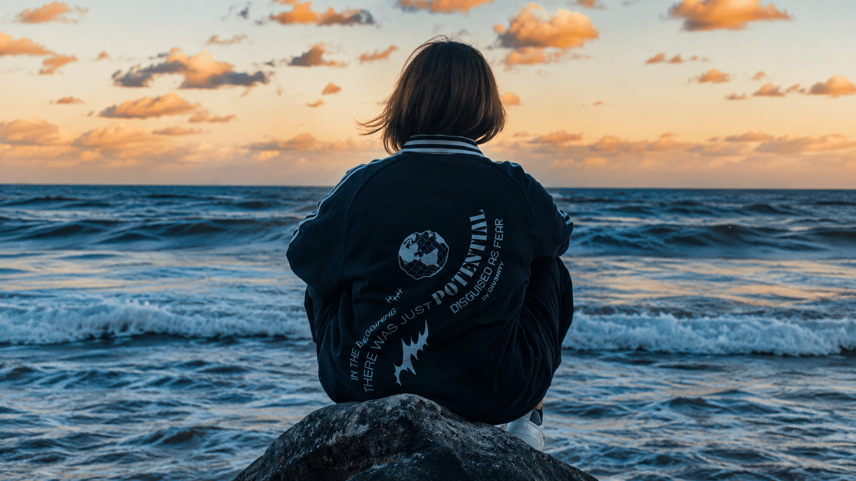 Mayte sentada sobre una roca frente al mar al atardecer con un jersey DIV3NITY